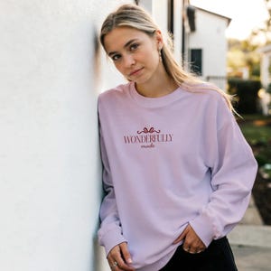 Può includere: Una felpa a girocollo a maniche lunghe di colore viola chiaro, caratterizzata da una piccola grafica rossa di un fiocco sopra la scritta "WONDERFULLY made" centrata sul petto. Il capo presenta una vestibilità rilassata con polsini a coste.