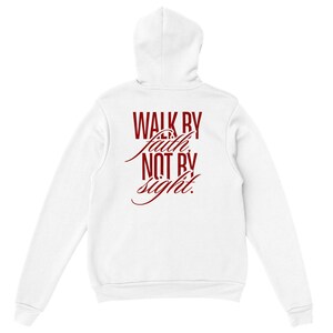 Può includere: Una felpa con cappuccio bianca con il testo "WALK BY faith, NOT BY sight." stampato in rosso scuro sul retro. Il design combina lettere in grassetto con un'elegante scrittura corsiva, centrate sulla parte superiore della schiena.