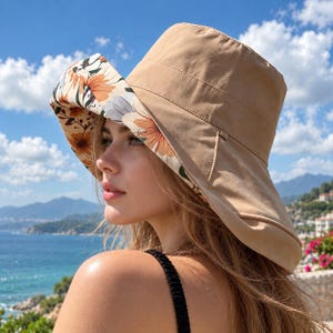 May include: A reversible bucket hat featuring a solid tan exterior and an interior lining with a floral pattern of orange, white, and grey flowers on a cream background. The hat has a wide, flexible brim and a structured crown design.