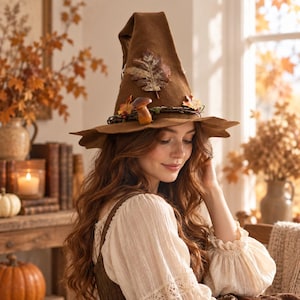 May include: A brown felt pointed hat featuring a wide brim with uneven edges. The hat is decorated with a large dried oak leaf, a small brown mushroom, and a thin twisted vine wrapped around the base, accented with tiny colourful autumn leaves.
