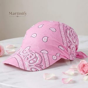 Może przedstawiać: Różowa czapka z daszkiem w biały wzór paisley typu bandana. Czapka posiada zakrzywiony daszek oraz materiałowe wiązanie z tyłu. Słowo Martinify jest wydrukowane czarnym tekstem nad czapką na jasnym tle.