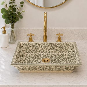 May include: A square ceramic vessel sink featuring a green and cream botanical leaf pattern. The sink includes a gold-toned drain and is paired with a matching gold-toned faucet and two cross-handle knobs mounted on a white stone countertop.