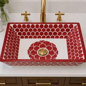 May include: A rectangular ceramic vessel sink featuring a white base with a repeating red scalloped pattern along the interior walls and a large red floral motif at the centre. The sink includes a circular brass drain plug centred in the basin.