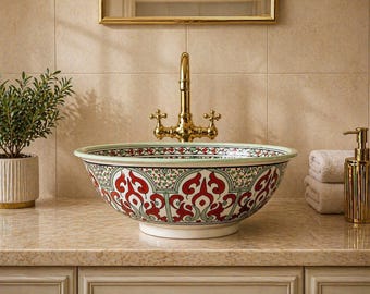 May include: A round ceramic vessel sink featuring a detailed pattern of red and green scrollwork on a white background. The basin has a wide, shallow bowl shape with a smooth rim and sits centered beneath a polished gold-toned metal faucet.