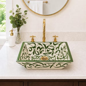 Puede incluir: Un lavabo cuadrado de cerámica con un patrón pintado a mano de enredaderas y volutas verdes sobre un fondo color crema. El lavabo incluye un desagüe de metal dorado y se combina con un grifo dorado a juego y dos mandos en forma de cruz.