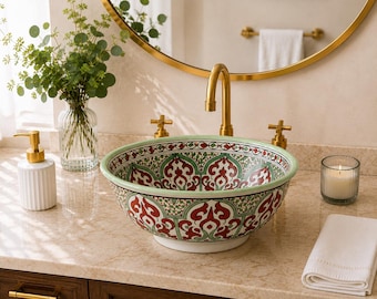 May include: A round ceramic vessel sink featuring a detailed red and green floral pattern on a white background with a light green rim. The sink is positioned on a beige countertop with a gold faucet, a white soap dispenser, and a white towel.