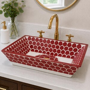 May include: A rectangular ceramic vessel sink featuring a deep red and white scalloped pattern on the interior walls and a central floral motif at the base. The sink is paired with a polished gold faucet and matching cross-handle hardware.