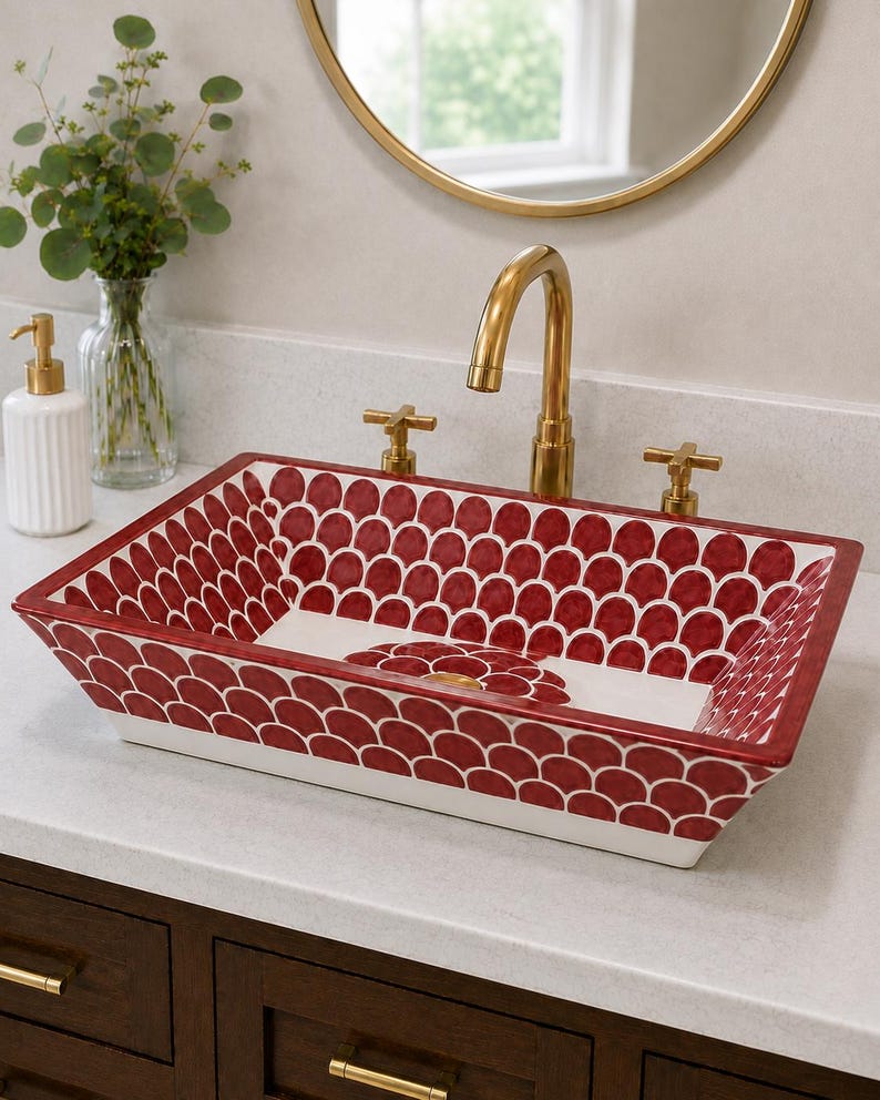 May include: A rectangular ceramic vessel sink featuring a deep red and white scalloped pattern on the interior walls and a central floral motif at the base. The sink is paired with a polished gold faucet and matching cross-handle hardware.