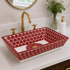 May include: A rectangular ceramic vessel sink featuring a repeating red and white scalloped pattern on the interior and exterior walls. The basin includes a central circular floral motif surrounding the drain and a gold-toned metal faucet fixture.