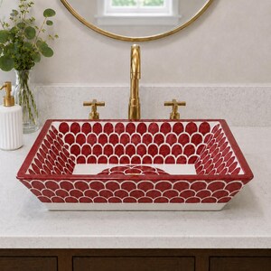 May include: A rectangular ceramic vessel sink featuring a repeating red and white scalloped pattern across its interior and exterior surfaces. The sink is paired with a tall, brushed gold faucet and two matching cross-handle knobs on a white countertop.