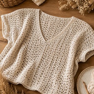 May include: A cream-colored short-sleeved top featuring an open-knit crochet pattern with a V-neckline. The garment is constructed with a textured, airy stitch design, short cap sleeves, and a straight hemline, showcasing a neutral off-white yarn.