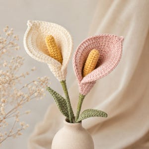 Puede incluir: Dos calas de ganchillo, una en color crema y otra en rosa empolvado, cada una con un espádice amarillo texturizado. Las flores tienen tallos y hojas verdes de ganchillo y están dispuestas juntas en un pequeño jarrón de cerámica neutro.