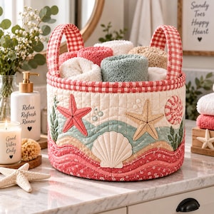 May include: A round quilted fabric storage basket featuring a coastal theme with appliqued starfish, seashells, and wavy patterns in shades of red, beige, and teal. The basket has two red and white gingham handles and contains rolled towels.