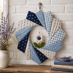 May include: A quilted fabric wreath featuring a starburst pattern of blue and white floral, gingham, and solid fabric segments. A central white fabric flower with a blue center and green felt leaves hangs from a string within the wreath center.