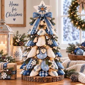 May include: A decorative tabletop Christmas tree constructed from layered fabric petals in shades of blue, white, and cream. The tree features patterns of snowflakes, plaid, and knit textures, topped with a star, a blue bow, and small wooden ornaments.