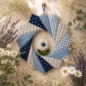 May include: A circular fabric wreath featuring a starburst pattern of blue and white quilted panels in floral, gingham, and solid prints. A small, three-dimensional white fabric flower with a blue center and green leaves hangs in the center.