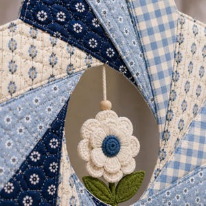 May include: A quilted star-shaped wall hanging featuring blue and white fabric patterns including gingham, floral, and geometric prints. A crocheted white flower with a blue center and green fabric leaves hangs from the center on a cord.