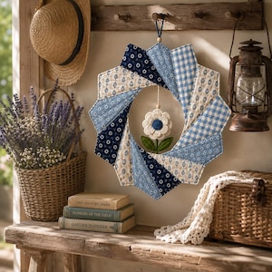 May include: A fabric wreath featuring a starburst pattern of blue and white quilted panels in floral, gingham, and solid prints. A small, cream-colored fabric flower with a blue center and green leaves hangs from the center of the circular opening.