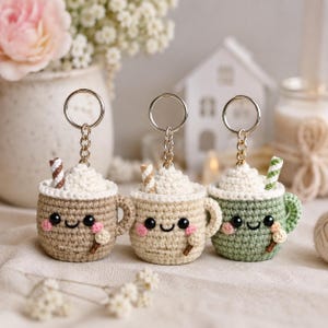 Può includere: Tre portachiavi a forma di tazza all'uncinetto con faccine sorridenti e guance rosa. Ogni tazza è sormontata da panna montata bianca all'uncinetto e una piccola cannuccia a righe. Le tazze sono marroni, beige e verdi, con un anello in metallo.