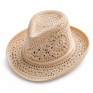 May include: A beige fedora hat crafted from woven straw material. The hat features a structured crown with a pinched top and a wide brim. The surface displays an intricate, open-work crochet pattern throughout, including a circular floral motif.