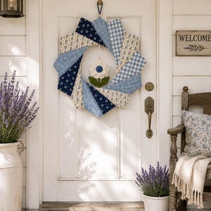 May include: A circular fabric wreath featuring a pinwheel design made of alternating blue and white patterned cotton segments. A small, white crocheted flower with a dark blue center and green leaves hangs from the center of the wreath.