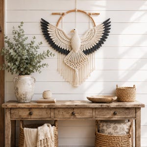 May include: A macrame wall hanging shaped like a bird with wings spread wide, featuring cream-colored cord for the body and head, and dark gray cord for the wing tips. The bird is suspended from a circular wooden hoop with hanging fringe below.