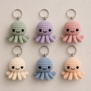 May include: Six crocheted octopus keychains arranged in two rows. Each keychain features a round head, eight small tentacles, and two black bead eyes. The octopuses are coloured in sage green, lavender, dusty pink, cream, light blue, and peach.
