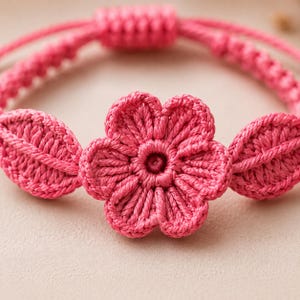 Könnte beinhalten: Ein leuchtend pinkfarbenes Häkelarmband mit einer zentralen fünfblättrigen Blume, die von zwei blattförmigen Akzenten flankiert wird. Das Band besteht aus geflochtener Kordel mit einem Schiebeknotenverschluss und zeigt feine Texturen.