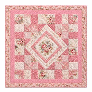 May include: A square quilt featuring a patchwork design with a central diamond motif. The fabric displays various shades of pink, cream, and white with floral patterns, including roses and small blossoms, finished with a quilted texture.