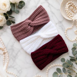 May include: Three crocheted headbands featuring a twisted front design. The headbands are colored in dusty rose, white, and deep burgundy. Each piece is crafted with a textured knit pattern and is arranged in a stack on a marble surface.