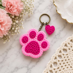 Peut inclure: Un porte-clés en crochet représentant une empreinte de patte dans deux tons de rose, rose clair et rose foncé. Le porte-clés comprend un anneau en métal couleur bronze et un petit cœur en crochet rose foncé uni attaché par une courte chaîne.