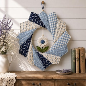 May include: A circular fabric wreath featuring a starburst pattern of quilted panels in shades of navy blue, light blue, and cream with floral and gingham prints. A small, crocheted white flower with a blue center hangs in the center.