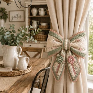 Puede incluir: Una cortina beige sujeta por un lazo de tela acolchada con un estampado floral en verde salvia y rosa empolvado. El lazo está decorado con pequeñas perlas blancas, bordes festoneados y dos adornos circulares de flores de tela.