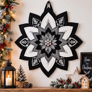 May include: A quilted fabric wall hanging shaped like a large star or flower with layered petals in black, white, and grey. The petals feature stitched leaf patterns and a centre cluster of small white spheres. It hangs from a fabric loop.