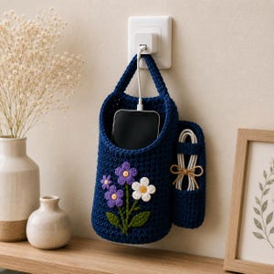 May include: A dark blue crocheted wall-hanging storage pouch featuring a floral motif with two purple flowers, one white flower, and green leaves. The pouch has a side pocket holding a white coiled charging cable and contains a black smartphone.
