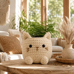 Puede incluir: Una maceta de ganchillo color crema con forma de gato, con dos orejas puntiagudas, dos pequeñas patas delanteras y una cara con ojos de cuentas negras y bigotes y nariz bordados en marrón, que contiene una pequeña planta de helecho verde.