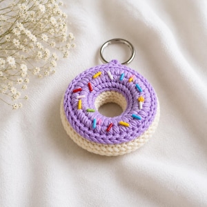 Könnte beinhalten: Ein gehäkelter Donut-Schlüsselanhänger mit einer cremefarbenen Basis und einer lavendelfarbenen Oberseite, die mit kleinen, bunten Garnstreuseln verziert ist. Ein silberfarbener Metallring ist an der Oberseite des textilen Objekts befestigt.