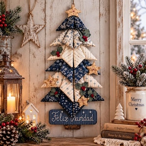 Puede incluir: Una decoración navideña de pared en forma de árbol hecha con triángulos de tela estampada en azul marino y crema, adornada con pequeñas estrellas de tela, hojas de acebo verdes y bayas rojas, con una base que lleva la inscripción Feliz Navidad.