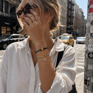 Puede incluir: Una pila de cinco pulseras en la muñeca, que incluye una hebra de cuentas, un dije dorado de mapa mundial, un símbolo de infinito, un dije circular con incrustaciones de cristales y un dije dorado con la palabra Love en letra cursiva.