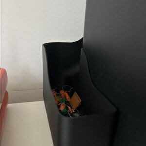 Könnte beinhalten: Ein schwarzer Kunststoff-Aufbewahrungsbehälter mit geschwungenem Seitenprofil, der an der Seite eines schwarzen elektronischen Geräts befestigt ist. Der Behälter enthält kleine Gegenstände, darunter ein quadratisches beiges Teil.