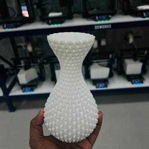 May include: A white 3D printed vase with a narrow neck and a bulbous body, featuring a repeating geometric pattern of small, raised triangular textures across the entire exterior surface, creating a tactile and structured appearance.