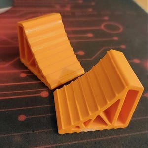 May include: Two bright orange, wedge-shaped wheel chocks featuring a stepped, ridged top surface and a hollow, triangular side profile with internal support structures. These objects are constructed with a visible layered texture.