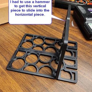 May include: A black 3D printed plastic stand with a grid of circular cutouts. A vertical piece is inserted into a horizontal base. A speech bubble contains the text: I had to use a hammer to get this vertical piece to slide into the horizontal piece.