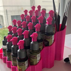 以下が含まれることがあります： 鮮やかなピンク色の階段状のプラスチック製オーガナイザー。ピンク色のスカル型キャップが付いた小さな塗料用ドロッパーボトルが多数収納されています。スタンドの高い側面コンパートメントには、数本の細い絵筆が立てられています。