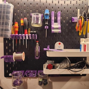 May include: A black pegboard organizer featuring purple 3D-printed mounts and hooks holding various tools, including screwdrivers, pliers, and a spool of solder. Two white shelves hold bottles and cables, while a clear plastic bin stores small parts.