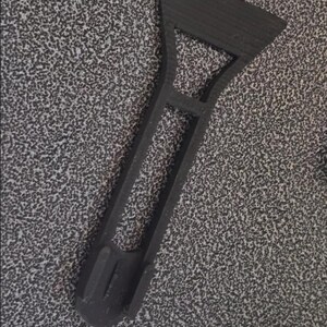 May include: A black plastic scraper tool featuring a wide, flat blade edge at the top, a central open rectangular handle section, and a rounded base with two small vertical tabs for gripping or attachment purposes, showing a textured surface finish.