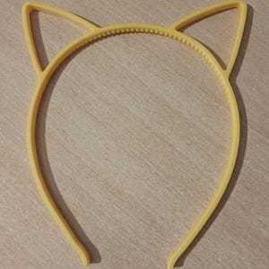 May include: A yellow plastic headband featuring two pointed cat ear shapes extending from the top. The inner band has a series of small, evenly spaced teeth designed for grip, and the entire structure is formed from a single piece of material.
