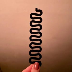 以下が含まれることがあります： 黒いプラスチック製のヘアスタイリングツール。長く狭い蛇行したジグザグの形状をしており、全長にわたって複数の丸みを帯びたループが並んでいます。髪の毛を隙間に通して複雑なヘアスタイルを作るのを補助するために設計されています。