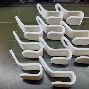 May include: Ten white 3D printed plastic hooks arranged in a grid. Each hook features a curved J-shape design with a flat mounting tab at the top containing a circular hole for attachment. The hooks have a smooth, matte finish and uniform shape.