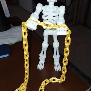 May include: A white articulated skeleton figurine standing upright and holding a long, continuous gold-colored plastic chain. The skeleton features a detailed skull, rib cage, and jointed limbs, while the chain consists of multiple interlocking oval links that drape downward from the skeleton's hands.